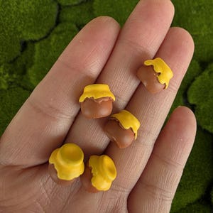 Pode incluir: Pequenos potes de mel castanhos com tampas amarelas são exibidos. Os pequenos potes estão dispostos em uma mão, com um fundo verde e musgoso. Os potes de mel têm um design charmoso e caprichoso.