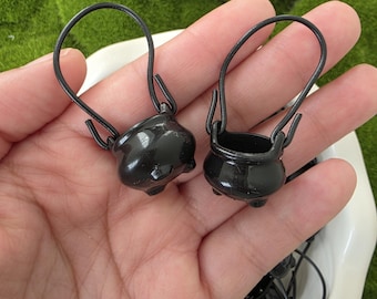 May include: Two small, glossy black cauldron-shaped charms, each featuring a rounded body with three short feet and a thin, arched metal handle attached to the sides. The charms have a smooth, reflective finish and a hollow, open top design.