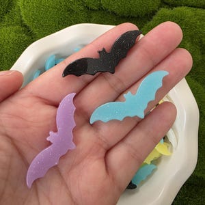 May include: Three glittery bat-shaped charms in black, light blue, and lavender. The charms are displayed in a white dish, with a green moss background. The bats have a sparkly finish, ideal for Halloween or craft projects.