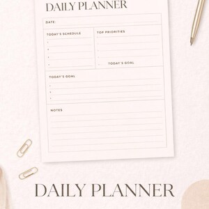 Puede incluir: Un planificador diario beige con el texto "DAILY PLANNER" en la parte superior e inferior. El planificador incluye secciones para la fecha, el horario, las principales prioridades y los objetivos. También se ve un bolígrafo dorado y clips.