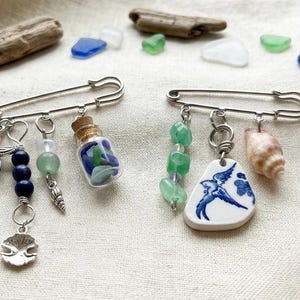May include: Two decorative safety pins with beach-themed charms. One pin features a starfish, blue beads, a shell, and a miniature bottle with blue and green glass. The other pin has green beads, a shell, and a porcelain pendant with a blue bird design.