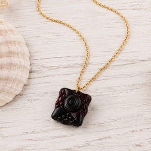 May include: A gold-toned metal chain necklace featuring a square-shaped pendant with a dark burgundy, textured surface and a raised, circular black center element. The pendant has a glossy finish and a distinct geometric pattern across its face.