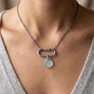 May include: A silver-toned metal chain necklace featuring an oval carabiner-style clasp as a central pendant. Attached to the bottom of the carabiner is a single, smooth, translucent light blue sea glass charm with a rounded, irregular shape.