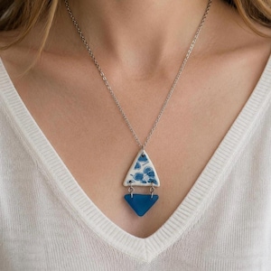 May include: A silver-toned chain necklace featuring a two-part pendant. The top piece is a white triangular ceramic charm with a blue floral pattern, connected by small metal rings to a solid, translucent dark blue triangular glass piece.