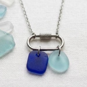 May include: A silver-toned necklace featuring a carabiner-style pendant with two sea glass charms. One charm is a square of deep blue glass, and the other is a light blue, oval-shaped piece. The necklace is displayed on a white surface.