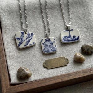 May include: Three porcelain pendant necklaces with silver chains. Each pendant features a unique blue and white design: birds, a pagoda, and a boat scene. The necklaces rest on a gray linen surface within a wooden tray.