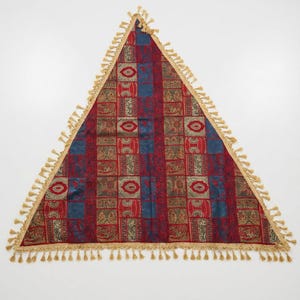 Puede incluir: Textil triangular con un diseño estampado en rojo, azul y dorado. La tela presenta un patrón geométrico repetitivo con borlas doradas en los bordes. Esta pieza decorativa podría usarse como mantel o tapiz.