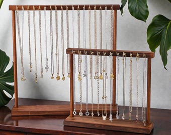 Puede incluir: Dos expositores de joyería de madera marrón oscuro con múltiples collares colgando. Los expositores tienen una base rectangular. Los collares son de diferentes longitudes y presentan diferentes colgantes, incluyendo llaves, estrellas y formas geométricas.