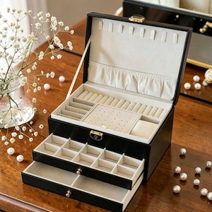 May include: Black jewelry box with multiple compartments and drawers. The interior is lined with a soft, light-colored fabric. The box has a gold-colored clasp and drawer pulls. The open box reveals various sections for storing jewelry.