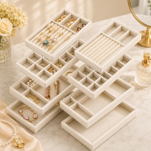 May include: A stack of off-white jewelry trays with various compartments and ring slots. The trays are filled with rings, bracelets, necklaces, and earrings. The jewelry is gold, silver, and pearl. The trays are on a marble surface.
