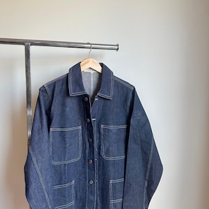 May include: A dark blue denim jacket with four front pockets and a button-up closure. The jacket has light-coloured stitching and is hanging on a wooden hanger. The jacket is displayed on a metal clothing rack against a neutral wall.