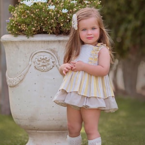 May include: A young child in a yellow and white striped dress with a layered skirt, white socks and sandals. The child has long blonde hair and a flower hair clip. A large white planter with flowers is in the background.