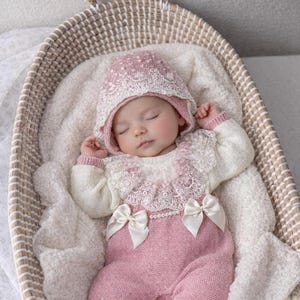 May include: A knitted baby outfit consisting of a dusty pink romper with two white satin bows at the waist and a matching bonnet. The garment features a white lace collar, long cream-colored sleeves, and a row of small white faux pearls at the waist.