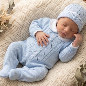 May include: A sleeping baby in a light blue knitted romper suit and matching hat with a white collar. The outfit has a cable knit design and pearl buttons. The baby lies on a cream-coloured, textured blanket.