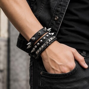 May include: A set of three black leather bracelets worn together. The top bracelet features large silver-toned metal spikes. The middle bracelet has a row of smooth silver-toned studs. The bottom bracelet displays a row of silver-toned pyramid studs.
