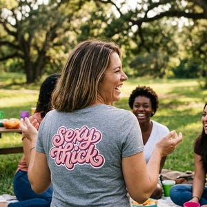 May include: A group of people laughing and enjoying a picnic outdoors. One person wears a gray t-shirt with the words "Sexy Thick" in a pink and white design. The scene is set in a park with trees and green grass.