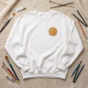 May include: A white crew neck sweatshirt featuring a small, embroidered smiling sun emblem on the left breast. The sun is yellow with an orange outline and a smiling face. Paintbrushes and art supplies are scattered around the sweatshirt.