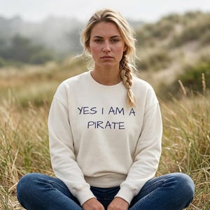 May include: Cream-colored sweatshirt with the text "YES I AM A PIRATE" in navy blue. The sweatshirt is a crew neck style. The person in the image is wearing blue jeans and has blonde hair in a braid.