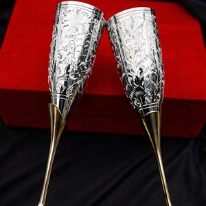May include: Two champagne flutes with intricate silver floral patterns on the bowls. The stems are gold, and the bases are a deep red. The flutes are set against a red box and a black background, creating a luxurious feel.