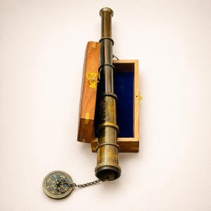 May include: A vintage brass telescope with a compass attached by a chain, displayed in an open wooden box with a blue velvet interior. The telescope is a dark brass color and has multiple sections. The box is a light brown color.