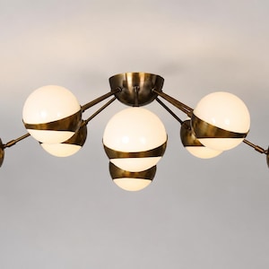 May include: A ceiling light fixture featuring a central brass canopy with multiple extending arms. Each arm holds a spherical white glass globe accented by a wide, horizontal brass band that wraps around the center of the orb, creating a layered look.