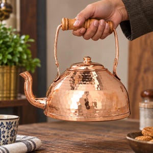 May include: A hammered copper tea kettle with a rounded body, a curved spout, and a cylindrical wooden handle. The kettle features a polished metallic finish with a textured surface and a matching lid with a small knob on top.
