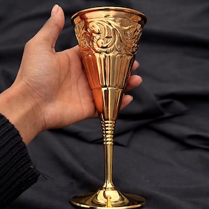 Könnte beinhalten: Ein goldfarbener Kelch mit aufwendigen Blumen- und Blattmustern. Der Kelch hat eine breite, geriffelte Schale und einen schlanken Stiel mit einem detaillierten Rippendesign. Der Fuß ist rund und der Kelch wird vor einem dunklen Hintergrund gehalten.