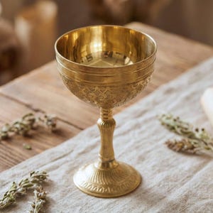 Puede incluir: Un cáliz dorado con un diseño en relieve ornamentado. El cáliz tiene un cuenco ancho, un tallo esbelto y una base circular. La superficie refleja la luz y parece ser de metal. El cáliz está sobre una superficie de madera.