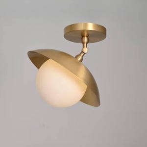 Op de afbeelding: Een messing plafondlamp met een ronde montageplaat, een verstelbaar draaigewricht en een brede, ondiepe, komvormige kap. Een bolvormige, matwitte glazen bol is in het midden van de messing kap geplaatst.