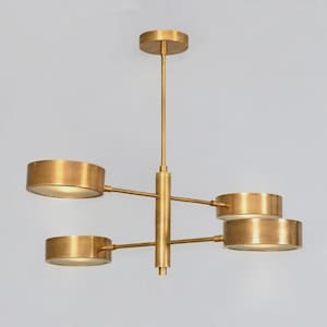 May include: A brass chandelier featuring a central vertical rod connected to a circular ceiling mount. Four horizontal arms extend from the center, each supporting a cylindrical drum shade with a metallic finish and a light-diffusing bottom panel.