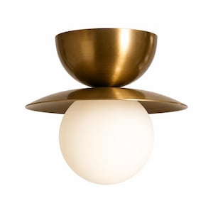 May include: A ceiling light fixture featuring a brushed brass finish with a bowl-shaped top, a flat circular disc in the middle, and a large white spherical glass globe at the bottom. The metal components have a smooth, metallic sheen.