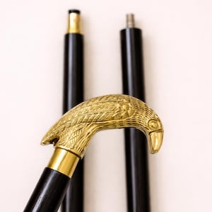 Puede incluir: Un bastón negro con un mango dorado en forma de cabeza de águila. El mango está intrincadamente detallado y el bastón tiene un eje negro. Dos ejes negros adicionales son visibles en el fondo.