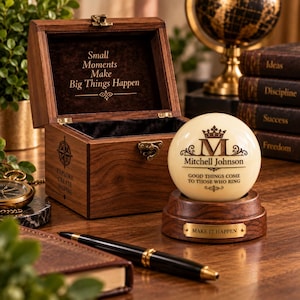 May include: A wooden box with a hinged lid featuring the text Small Moments Make Big Things Happen, alongside a cream-coloured sphere on a wooden base engraved with the letter M, the name Mitchell Johnson, and Good Things Come To Those Who Ring.