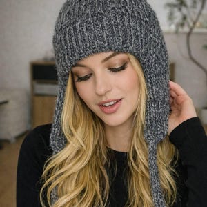 May include: A gray knit hat with ear flaps and a pom-pom. The hat is made of a thick, textured yarn and is designed to keep the wearer warm. The hat is a neutral color and can be worn with a variety of outfits.