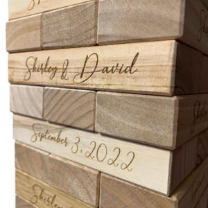May include: Close-up of a wooden block game with engraved text. The blocks are light brown and feature the names "Shirley & David" and the date "September 3, 2022" in elegant script. The game is stacked, showing multiple layers.