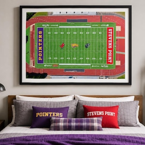 May include: A framed aerial view of a football field with the words "Stevens Point" and "Pointers" visible. The image is above a bed with pillows and a purple comforter. A pennant and a lamp are on the left.