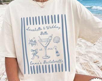 Puede incluir: Una camiseta de manga corta color marfil con un diseño gráfico azul enmarcado por rayas verticales. El estampado incluye una copa de martini, una concha, un bikini, una campana y una botella de champán con el texto Seashells & Wedding Bells est. 2026 Emma's Bachelorette.