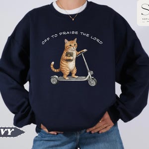 Puede incluir: Sudadera azul marino con un gato atigrado naranja montado en un patinete y sosteniendo un libro. El texto "OFF TO PRAISE THE LORD" est&aacute; arqueado sobre el gato. La palabra "NAVY" est&aacute; en la parte inferior izquierda.