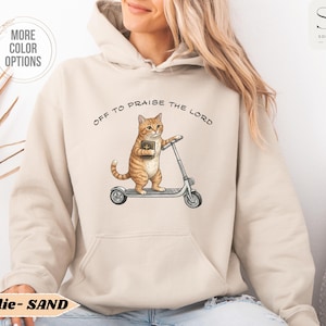 Puede incluir: Sudadera con capucha de color arena con un gato de dibujos animados montado en un patinete y sosteniendo un libro. El texto "OFF TO PRAISE THE LORD" est&aacute; impreso encima del gato. La sudadera tiene un bolsillo delantero y una capucha.
