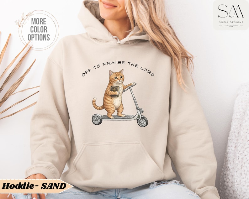 Puede incluir: Sudadera con capucha de color arena con un gato de dibujos animados montado en un patinete y sosteniendo un libro. El texto "OFF TO PRAISE THE LORD" est&aacute; impreso encima del gato. La sudadera tiene un bolsillo delantero y una capucha.