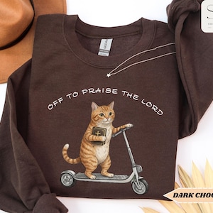Puede incluir: Sudadera marr&oacute;n chocolate oscuro con el texto "OFF TO PRAISE THE LORD". El gr&aacute;fico presenta un gato atigrado sosteniendo un libro y montando un scooter. Un sombrero marr&oacute;n es visible en la esquina superior izquierda.