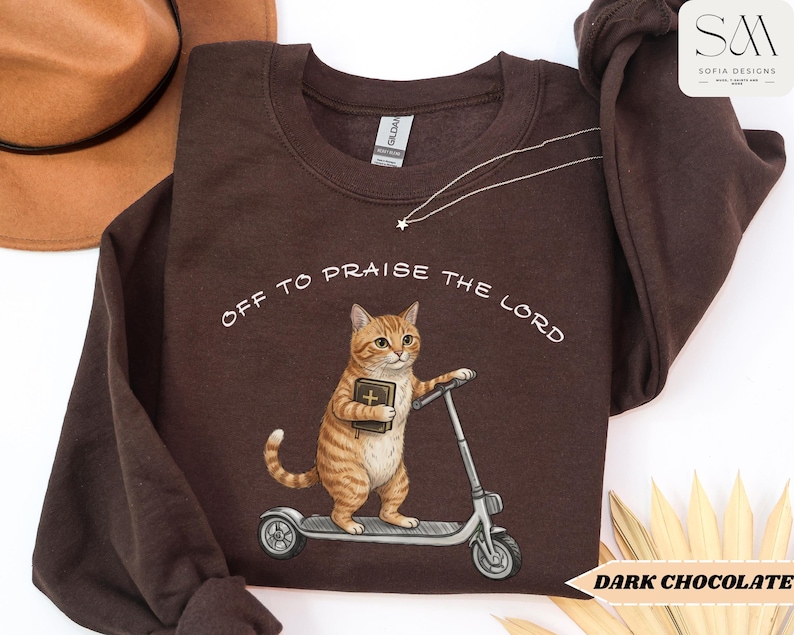 Puede incluir: Sudadera marr&oacute;n chocolate oscuro con el texto "OFF TO PRAISE THE LORD". El gr&aacute;fico presenta un gato atigrado sosteniendo un libro y montando un scooter. Un sombrero marr&oacute;n es visible en la esquina superior izquierda.
