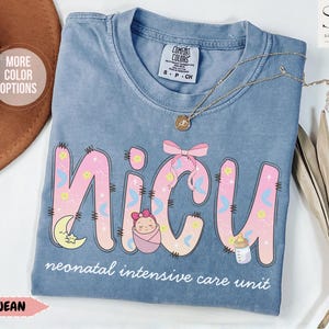 May include: Blue jean Comfort Colors t-shirt with the word "NICU" in pink and yellow, decorated with a moon, baby, bow, and bottle. The text "neonatal intensive care unit" is below the word "NICU". A gold necklace is draped on the shirt.