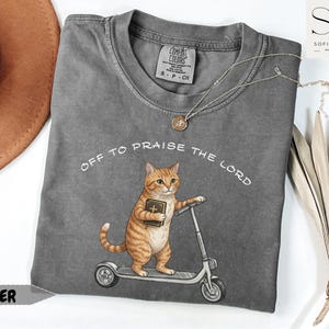 Puede incluir: Una camiseta gris Comfort Colors con el texto "Off to Praise the Lord". La camiseta presenta un gato de dibujos animados montado en un scooter mientras sostiene un libro. Un collar plateado con un colgante est&aacute; en la camiseta.