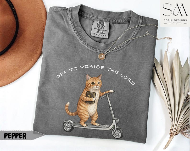 Puede incluir: Una camiseta gris Comfort Colors con el texto "Off to Praise the Lord". La camiseta presenta un gato de dibujos animados montado en un scooter mientras sostiene un libro. Un collar plateado con un colgante est&aacute; en la camiseta.