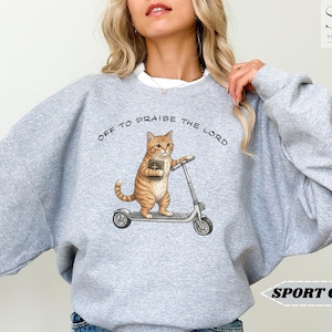 Puede incluir: Sudadera gris deportiva con un gato de dibujos animados en un patinete y un libro. El texto "OFF TO PRAISE THE LORD" est&aacute; sobre el gato. La sudadera es de color gris claro.