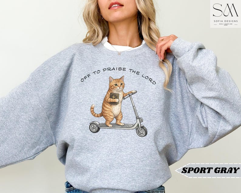 Puede incluir: Sudadera gris deportiva con un gato de dibujos animados en un patinete y un libro. El texto "OFF TO PRAISE THE LORD" est&aacute; sobre el gato. La sudadera es de color gris claro.