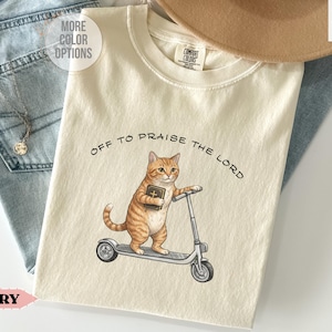 Puede incluir: Camiseta de color marfil con una ilustraci&oacute;n de un gato atigrado montando un patinete y sosteniendo un libro. El texto "OFF TO PRAISE THE LORD" est&aacute; impreso encima del gato. La camiseta es de material suave.