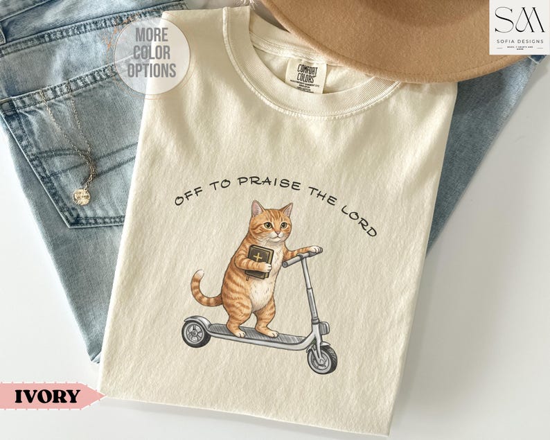 Puede incluir: Camiseta de color marfil con una ilustraci&oacute;n de un gato atigrado montando un patinete y sosteniendo un libro. El texto "OFF TO PRAISE THE LORD" est&aacute; impreso encima del gato. La camiseta es de material suave.