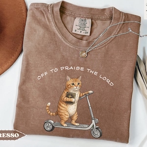 Puede incluir: Camiseta color espresso con el texto "Off to Praise the Lord". La camiseta presenta un gato de dibujos animados montado en un scooter y sosteniendo un libro. La camiseta tiene cuello redondo y mangas cortas. La etiqueta de la marca es visible.